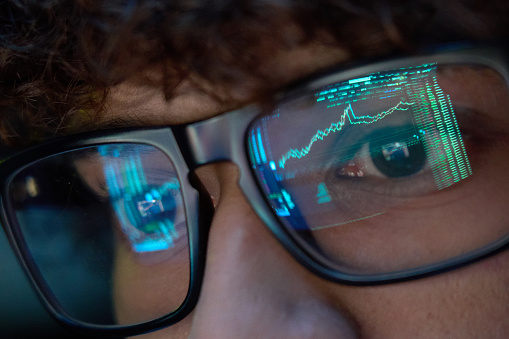Young indian business man trader wearing glasses looking at computer screen with trading charts reflecting in eyeglasses watching stock trading market financial data growth concept, close up.