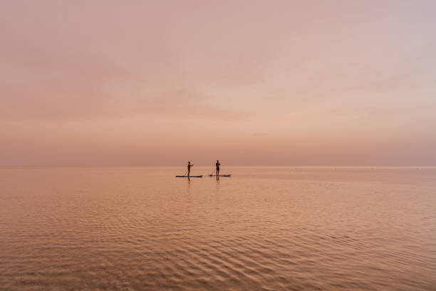 Photo of a young couple enjoying the beautiful sunset over the ocean, as they are stand up paddleboarding together.