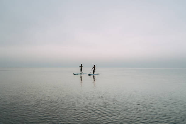 Photo of a young couple enjoying the beautiful sunset over the ocean, as they are stand up paddleboarding together.