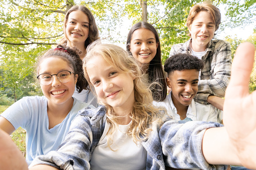 A small group of culturally diverse teenagers sit outside posing for a selfie. They are each dressed casually and sitting in close to one another as they smile.  The blond girl in front is holding out the camera for the group.