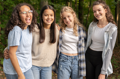 A small group of four culturally diverse teenage girls pose for a casual portrait outside. They are standing in a line, shoulder-to-shoulder, with their arms around one another as they smile.