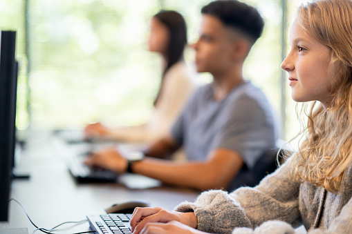 A small group of three Elementary students sit at a long desk in front of their own individual computers..  They are each dressed casually and typing away during the computer lab as they focus on the assignment.