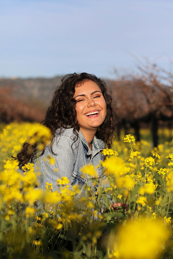 A girl sits in a vibrant yellow flower field while she smiles at the camera. Flowers are surrounding her with blue skies in the background.