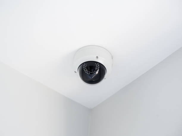 istock Digital security eye. A white round indoor CCTV surveillance camera monitoring security on ceiling in the room corner inside the white clean building. 1386610516