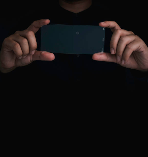 istock Future transparent glass phone Technology. Mockup blank horizontal screen futuristic super slim transparent smartphone in person's hands, taking photograph action, on black background with copy space. 1386610525