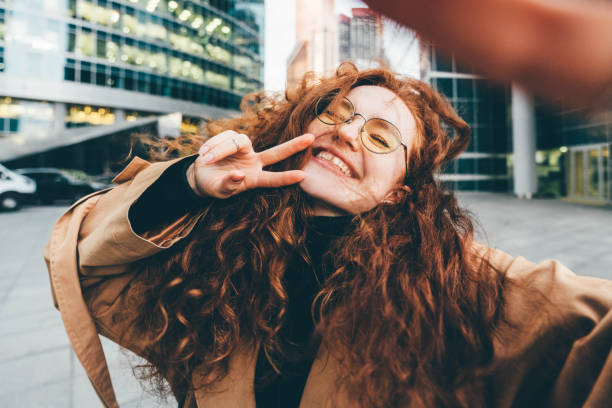 Curly haired young woman wearing fashion clothes with light makeup in glasses makes selfie posing for camera against modern skyscrapers in evening stock photo