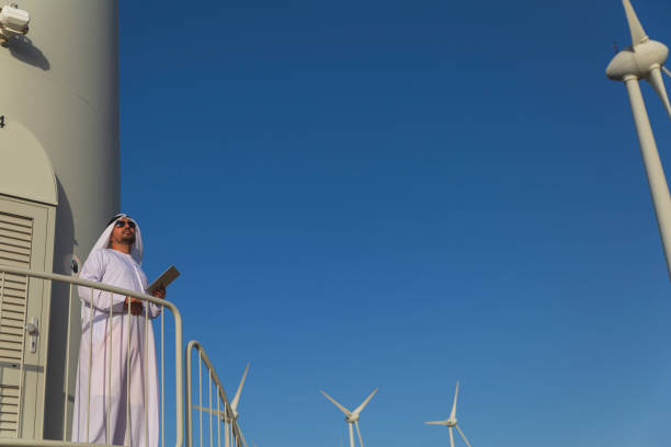 Arab Sheikh business man with traditional Emirati clothes looking for the energy industry and standing beside a wind turbines farm power station.