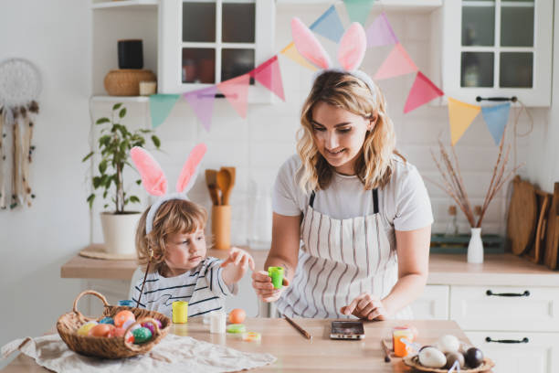 charming woman and her two-year-old son in bunny ears paint Easter eggs with paints sitting at the table on the kitchen stock photo