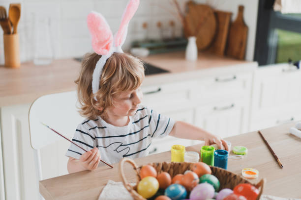 istock Cute two-year-old boy in bunny ears paint Easter eggs with paints sitting at the table on the kitchen 1386628065