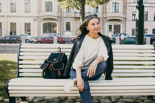 People, lifestyle, age, urban street style and fashion concept. Outdoor image of gorgeous attractive woman pensioner in trendy clothes waiting for friend on bench in park, enjoying nice sunny day