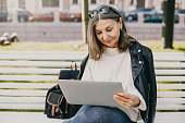 Successful fashionable mature retired woman wearing black leather jacket and blue skinny jeans sitting on bench with portable computer on her lap, having video conference call with her old friend