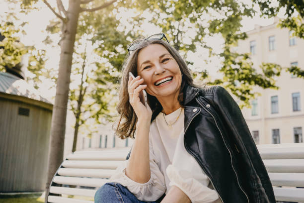 low angle view of cheerful beautiful european female in her fifties enjoying nice summer weather outdoors, receiving good positive news while having phone conversation, smiling broadly, laughing - casaco de couro imagens e fotografias de stock