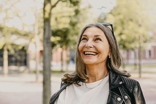 Cheerful overjoyed middle aged woman wearing sunglasses on her head and black leather jacket over white blouse enjoying peaceful beautiful morning while waking in park, looking up with broad smile