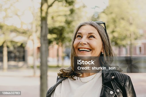istock Cheerful overjoyed middle aged woman wearing sunglasses on her head and black leather jacket over white blouse enjoying peaceful beautiful morning while waking in park, looking up with broad smile 1386632783