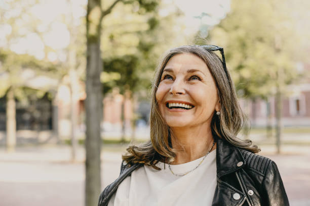 mulher alegre de meia-idade alegre usando óculos escuros na cabeça e jaqueta de couro preta sobre blusa branca desfrutando de manhã tranquila e bonita enquanto acordava no parque, olhando para cima com um sorriso largo - women smiling mature adult portrait - fotografias e filmes do acervo