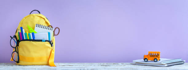 istock Full yellow school backpack with stationery and notes, pens, glasses on table on purple background. Banner. 1386638631