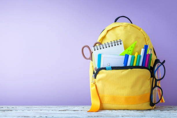 istock Full yellow school backpack with stationery and notes, pens, glasses on table on purple background. Concept back to school. 1386638647