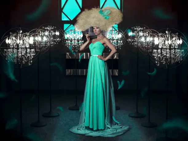 Photo of Beautiful and elegant woman wearing a turquoise gown with a big blonde wig, her head in a round cage, surrounded with lights and flying feathers, in a dark room
