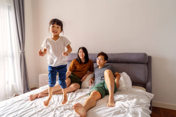 Happy family in bedroom and young girl child jumping on bed. stock photo