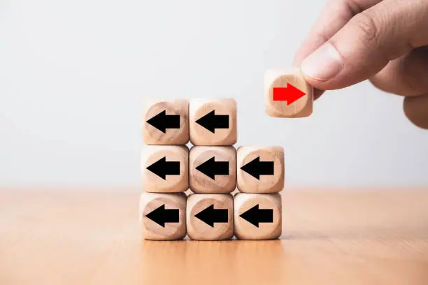 Photo of Hand holding red arrow on wooden move on different direction with black arrow on cube for business and technology disruption or unique concept.
