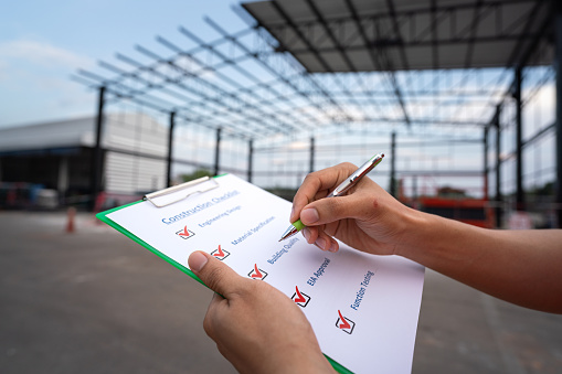 Action of an engineer is checking construction quality on the paperwork with warehouse building structure as blurred background. Industrial expertise working concept, close-up and selective focus.