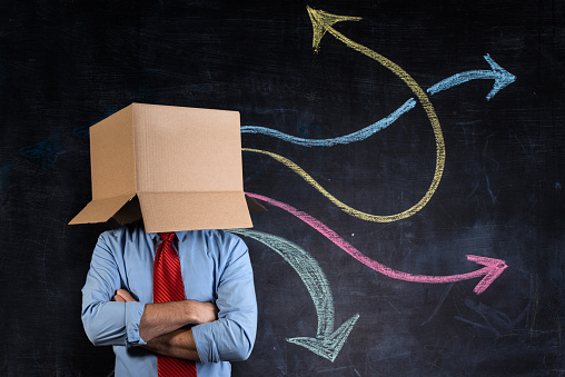 Businessman hiding his head in a cardboard box with crossed arms in front a blackboard showing many multicoloured arrows going in different directions