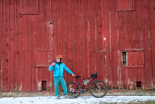 senior male cyclist is taking a break from winter riding on a gravel bike at old red barn in Colorado coutryside