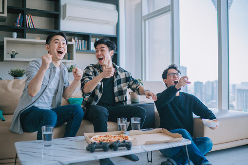 Asian Chinese young man shouting and cheering for his team support while watching sports on tv from home with his friends. Celebrate Victory when Sports Team Wins Championship. Friends Cheer, Shout