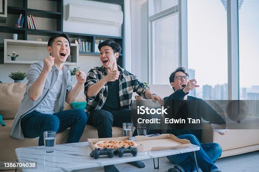 istock Asian Chinese young man shouting and cheering for his team support while watching sports on tv from home with his friends. Celebrate Victory when Sports Team Wins Championship. Friends Cheer, Shout 1386704038