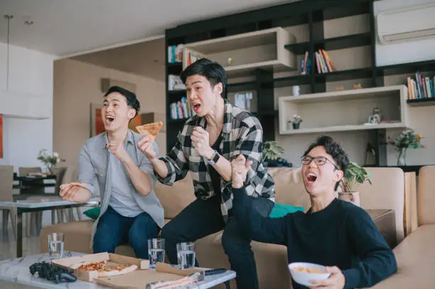 Photo of Asian Chinese young man shouting and cheering for his team support while watching sports on tv from home with his friends. Celebrate Victory when Sports Team Wins Championship. Friends Cheer, Shout