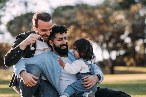 männliche schwule familie umarmt und spielt mit ihrer kleinen tochter im park. - homosexual stock-fotos und bilder