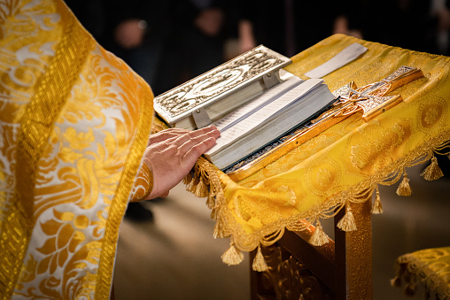 the hand prayer of the orthodox priest lies on the bible, in the foreground the cross and the service of the Lord takes place in the church