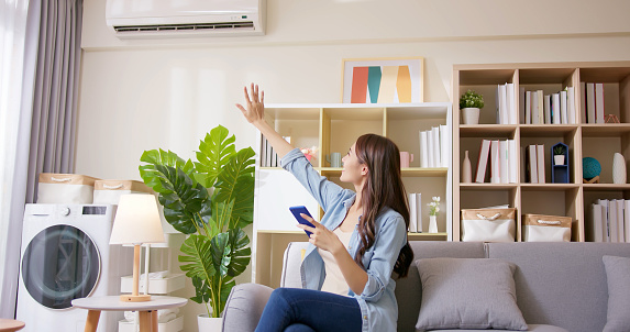 IOT smart home system concept - asian young woman using app on mobile phone to turn on air conditioner in living room