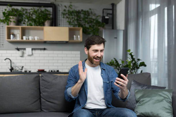 istock Men at home talking on a video call on the phone, using the application to communicate, sitting on the couch and waving his hand at the camera 1386725722