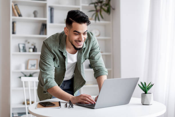 istock Smiling Handsome Arab Man Using Laptop At Home Office, Standing Near Desk 1386730594