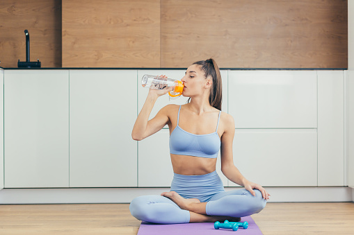 Portrait of a beautiful young fitness woman sitting on a mat in the lotus position and drinking water, juice after workout, yoga, at home, looking at camera, smiling