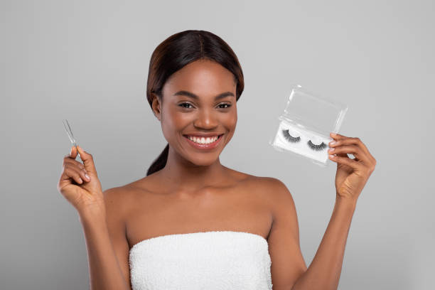 Black woman wrapped in towel holding pack of false eyelashes and tweezers Smiling beautiful black woman wrapped in towel holding pack of false eyelashes and tweezers while posing on grey background, happy young african american female ready to apply artificial eye lashes false lashes stock pictures, royalty-free photos & images