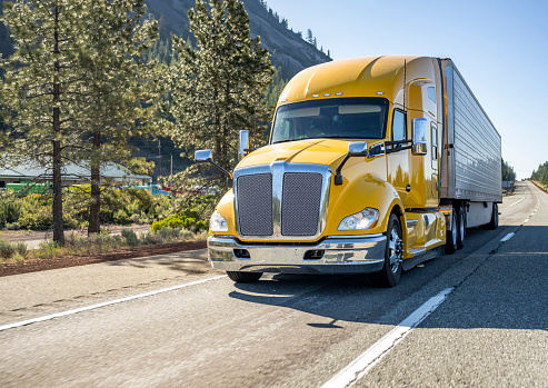 Classic long haul big rig yellow semi truck tractor with truck driver cab sleeping compartment transporting cargo in refrigerator semi trailer running on the highway road with rest area on the side