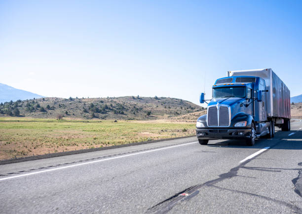 Classic long haul big rig blue semi truck with truck driver cab sleeping compartment transporting cargo in dry van semi trailer running on the road with hills and field on the side in California