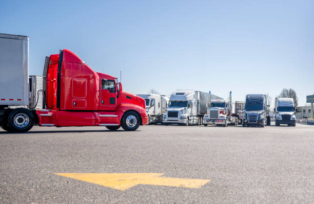 Bright red big rig semi truck with extended cab transporting cargo in dry van semi trailer driving on the truck stop parking lot with direction of movement arrow and row of another semi trucks stock photo