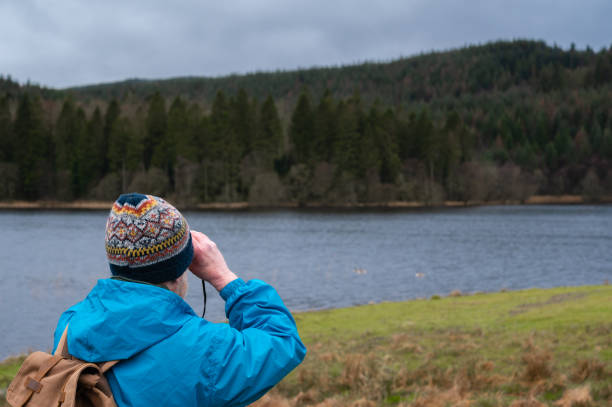 Senior man bird watching at a remote rural Scottish lake on an overcast and cold spring morning