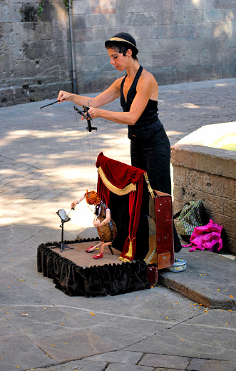 This is a photograph from 2013 of a street performer in Barcelona, Spain. Here, the female puppeteer manipulates various strings in order to create human-like movements in the puppet. Furthermore, the puppet's stage looks to be portable and appears as though it can be folded within some kind of a traveling suitcase.