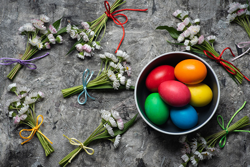 Top view of a group of colorful Easter eggs and spring flowers in a bowl on a rustic gray background.