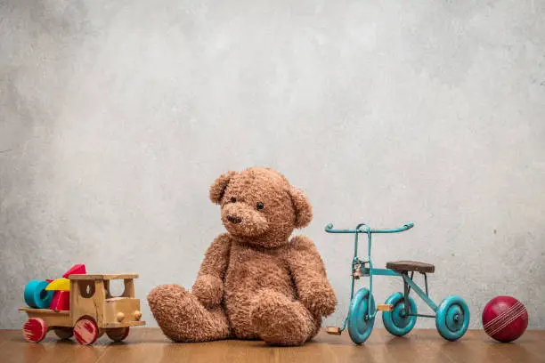 Photo of Retro plush Teddy Bear, old toy trike bicycle, obsolete wooden truck with construction blocks and leather ball front concrete textured wall background. Vintage style filtered photo