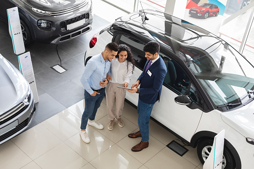 Top view of wealthy middle eastern young couple having conversation with male sales assistant at luxury auto showroom, checking nice white car and smiling, full length photo, copy space
