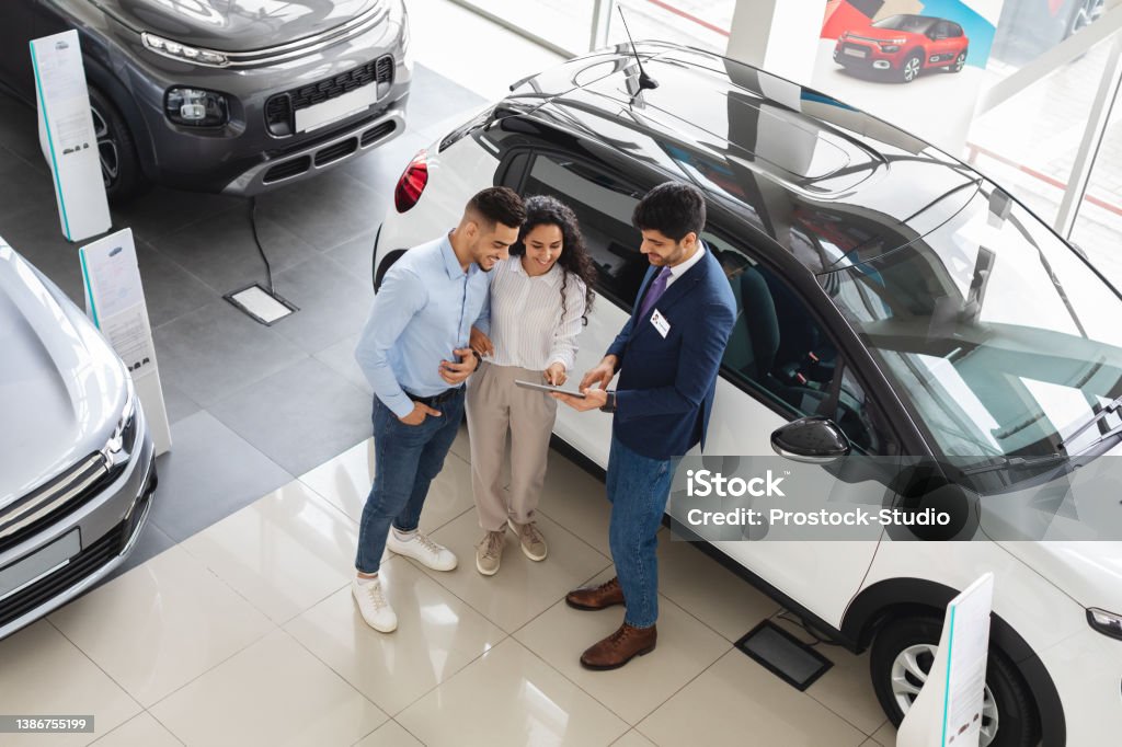 Customers having conversation with sales assistant, buying car, view above Top view of wealthy middle eastern young couple having conversation with male sales assistant at luxury auto showroom, checking nice white car and smiling, full length photo, copy space Car Dealership Stock Photo