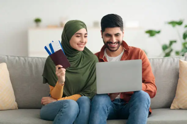 Cheerful Middle Eastern Spouses Holding Boarding Pass Using Laptop Booking Tickets Online Planning Vacation Together Sitting On Couch At Home. Technology And Transportation Concept