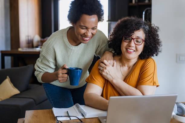 Cheerful gay couple looking at the laptop Portrait of two lesbian women using laptop and drinking coffee. They are having fun. homosexual couple stock pictures, royalty-free photos & images