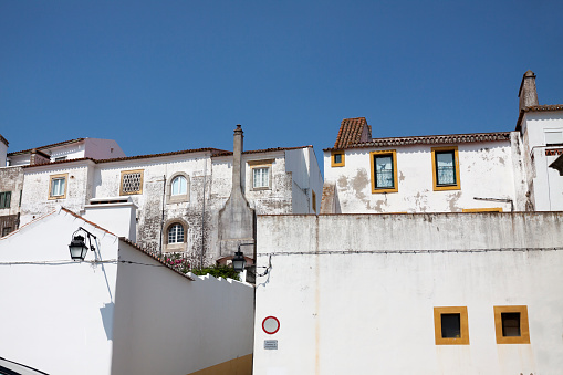 Whitewashed buildings in the city of Evora in Portugal. Scene dominated by large whitewashed walls in the foreground with two very small square windows with yellow frames in the wall to the right. Clear blue sky overhead with no clouds.