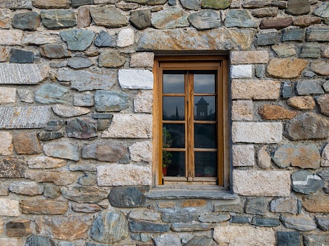 Old wooden window in a Hand-made natural stone building The wall is made of different size and shape stones. Flowers and flower pots are visible through the window. Reflection of the church.